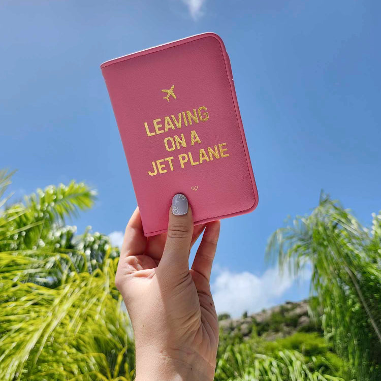 Leaving On A Jet Plan Passport Wallet in pink with gold foil Leaving On A Jet Plane text held up against the sky