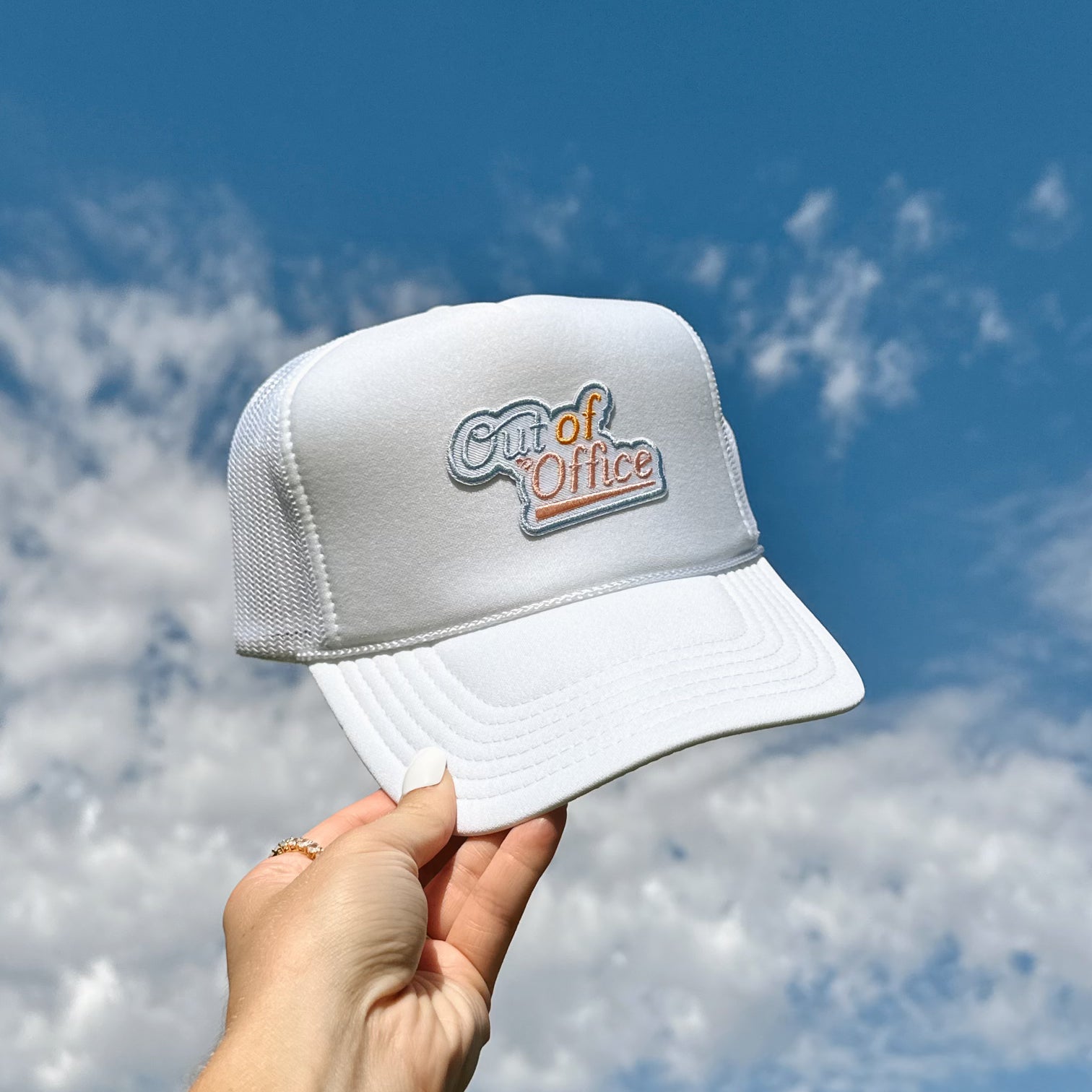 Out of Office Trucker Hat, white foam trucker cap with blue and orange Out of Office patch held up against blue sky