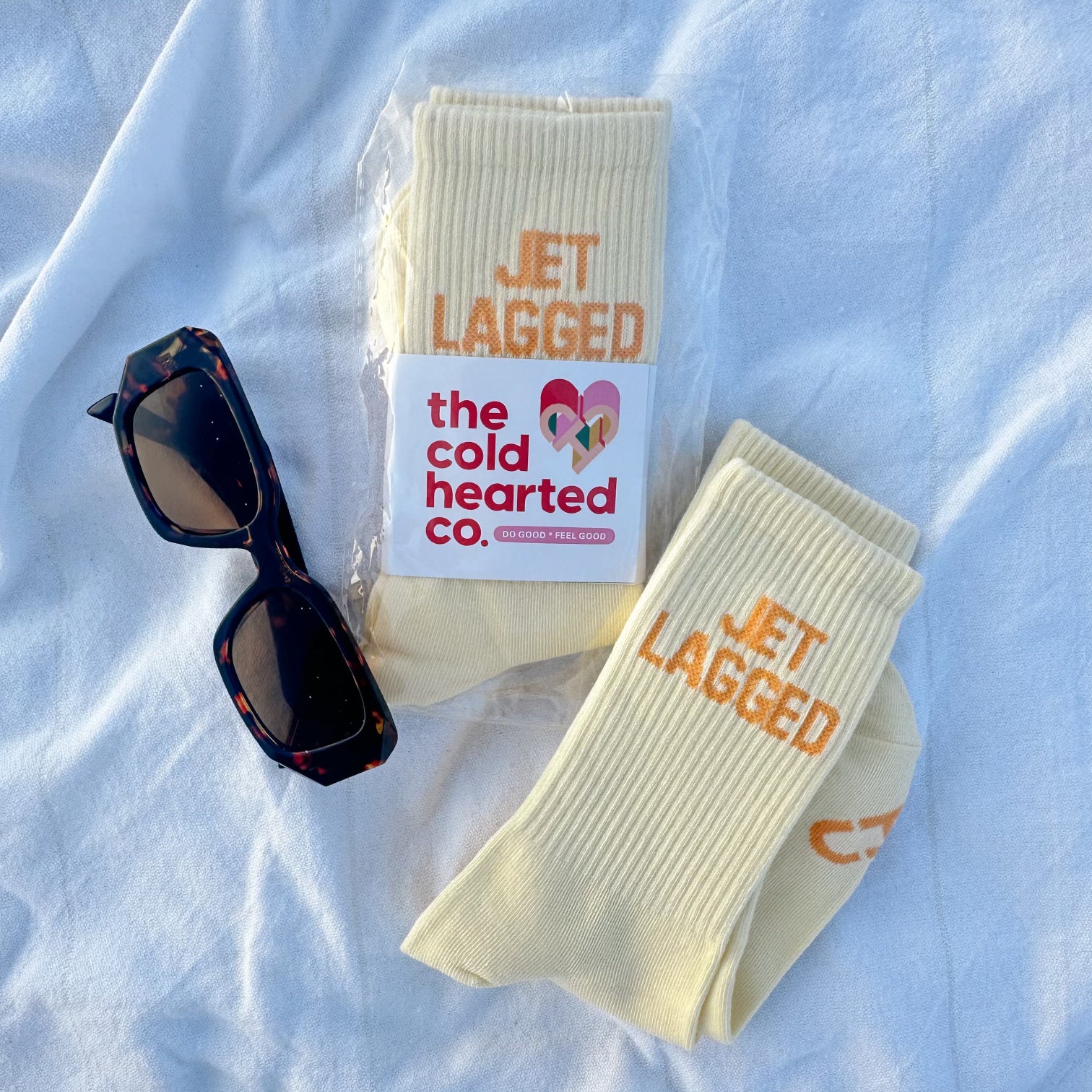 Jet Lagged Socks, pale yellow crew socks with orange Jet Lagged text and brand band, flat lay with tortoise sunglasses