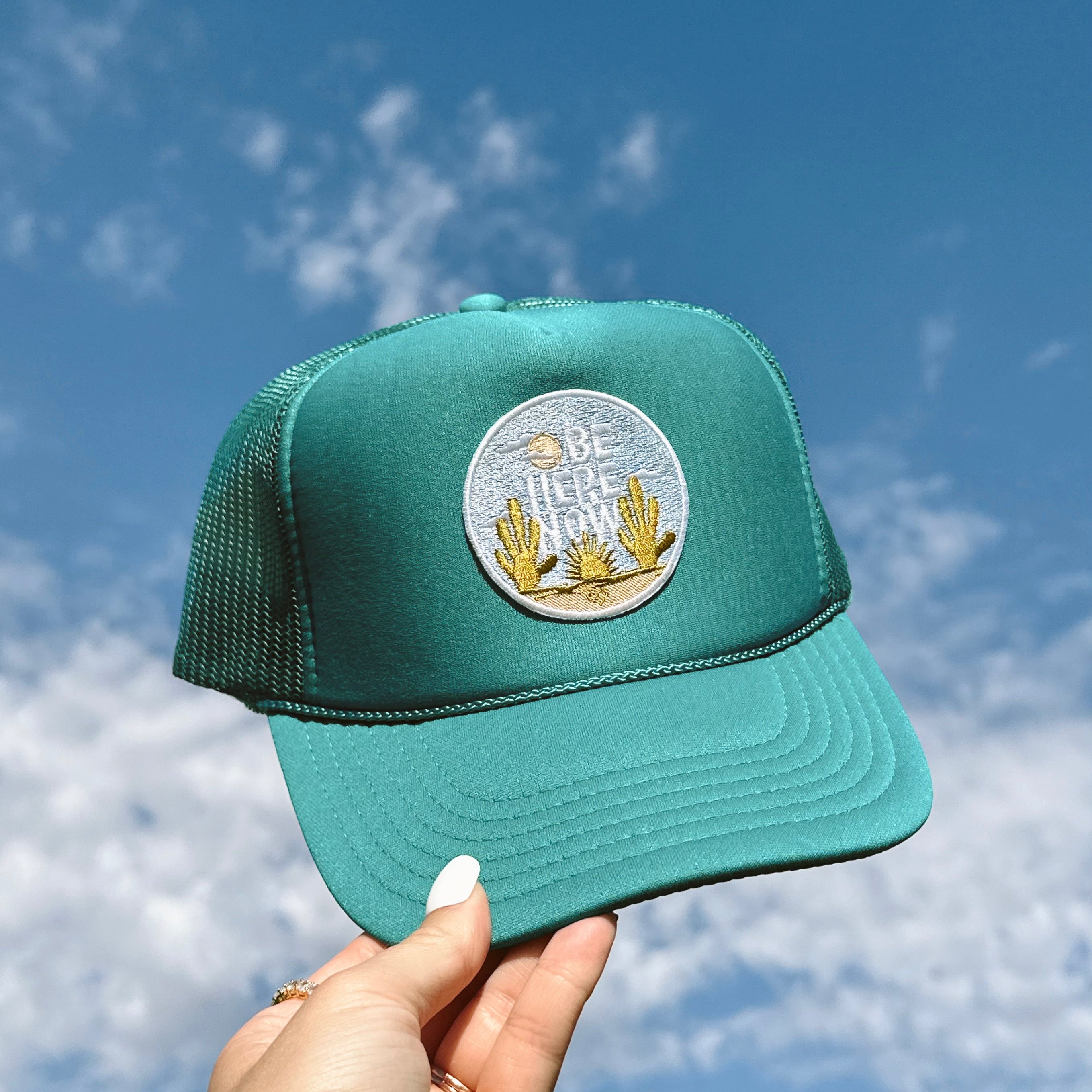 Be Here Now Trucker Hat, teal foam trucker hat with a round desert cactus patch held up against a blue sky