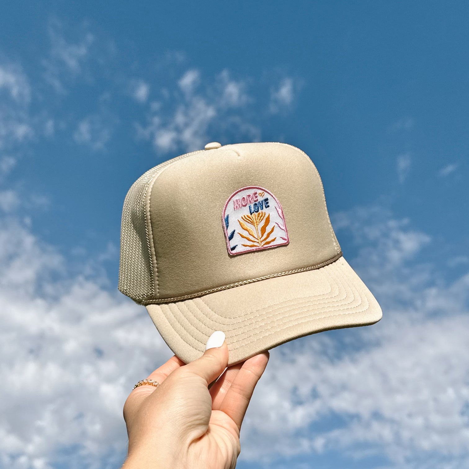 More Love Trucker Hat, tan foam trucker hat with an arched More Love plant patch held up against a blue sky