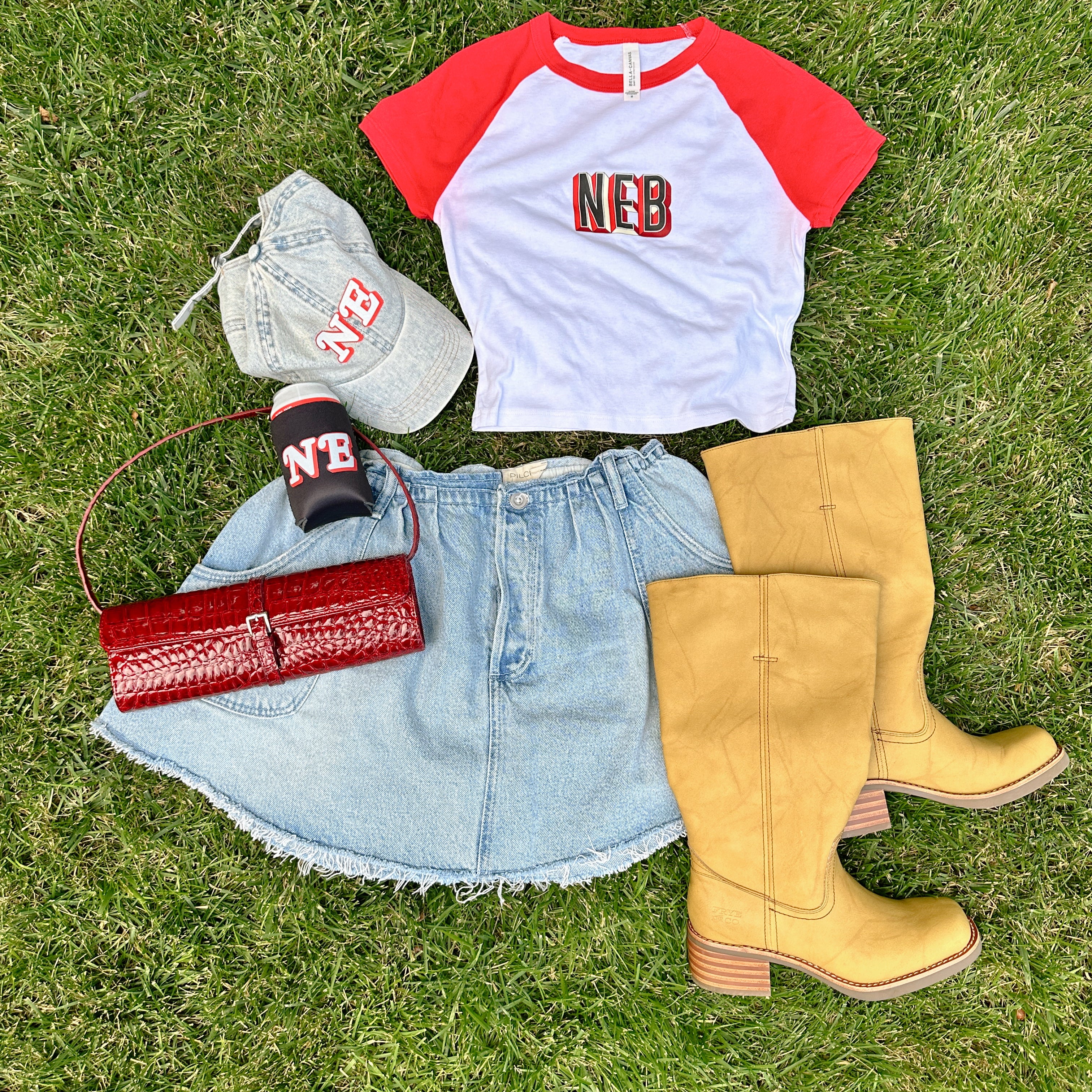 NE Denim Hat in a flat lay on grass with the NEB raglan baby tee, NE black koozie, denim skirt and tan boots