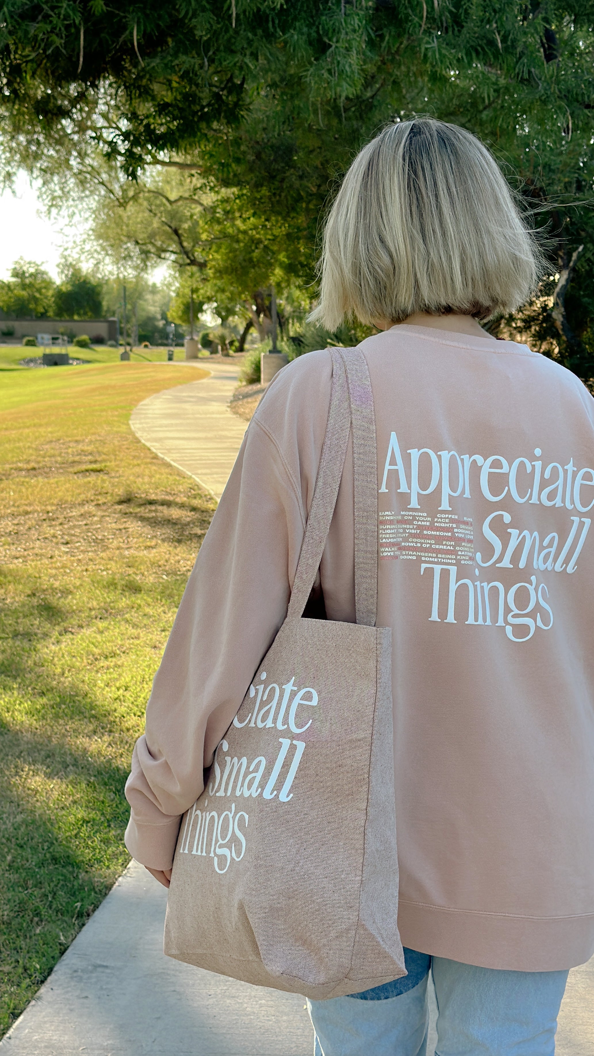 Appreciate Small Things Crew, back view of peach crewneck on a model walking a park path with a matching tote