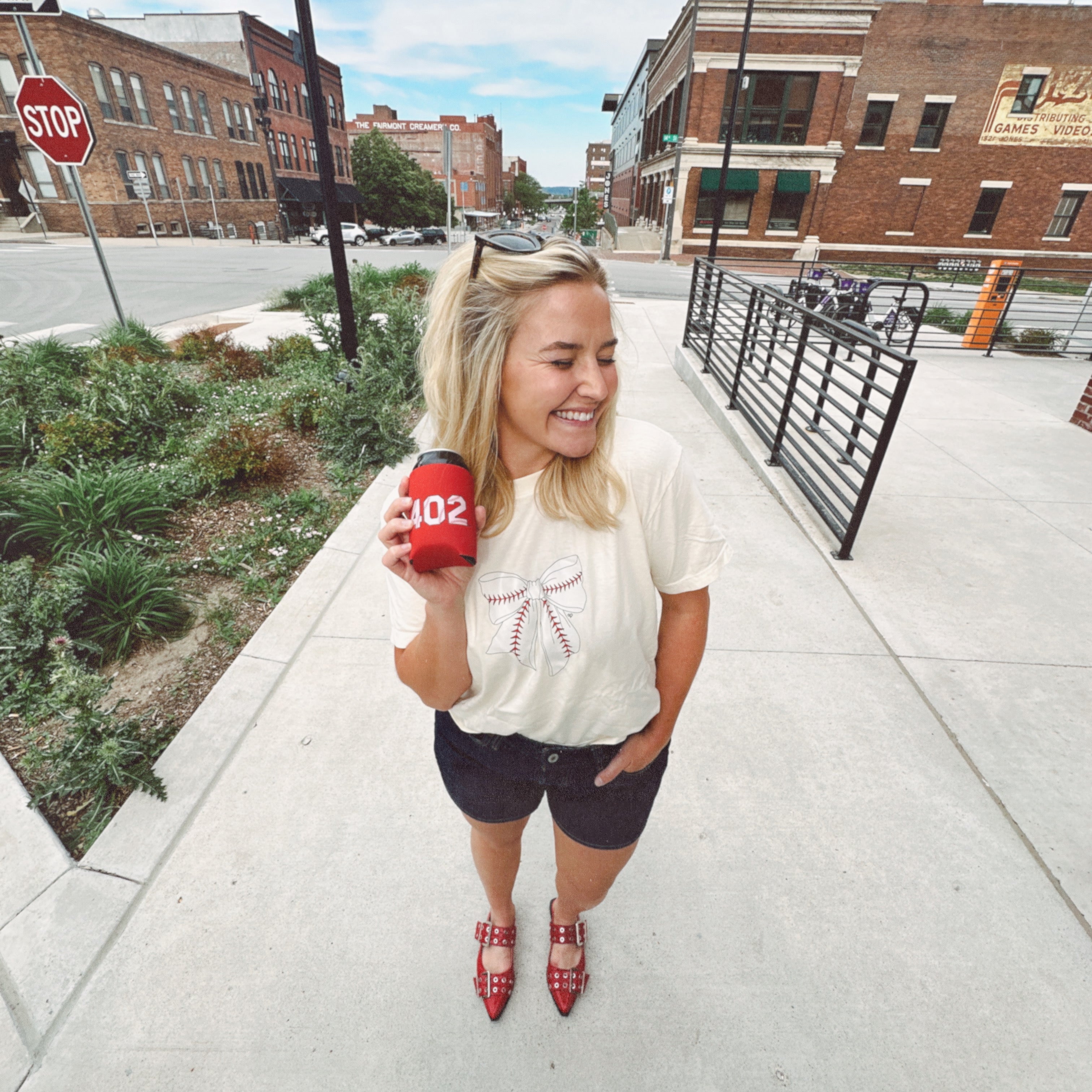 Baseball Bow Tee worn by a smiling model holding a red 402 koozie on a brick lined street in Omaha