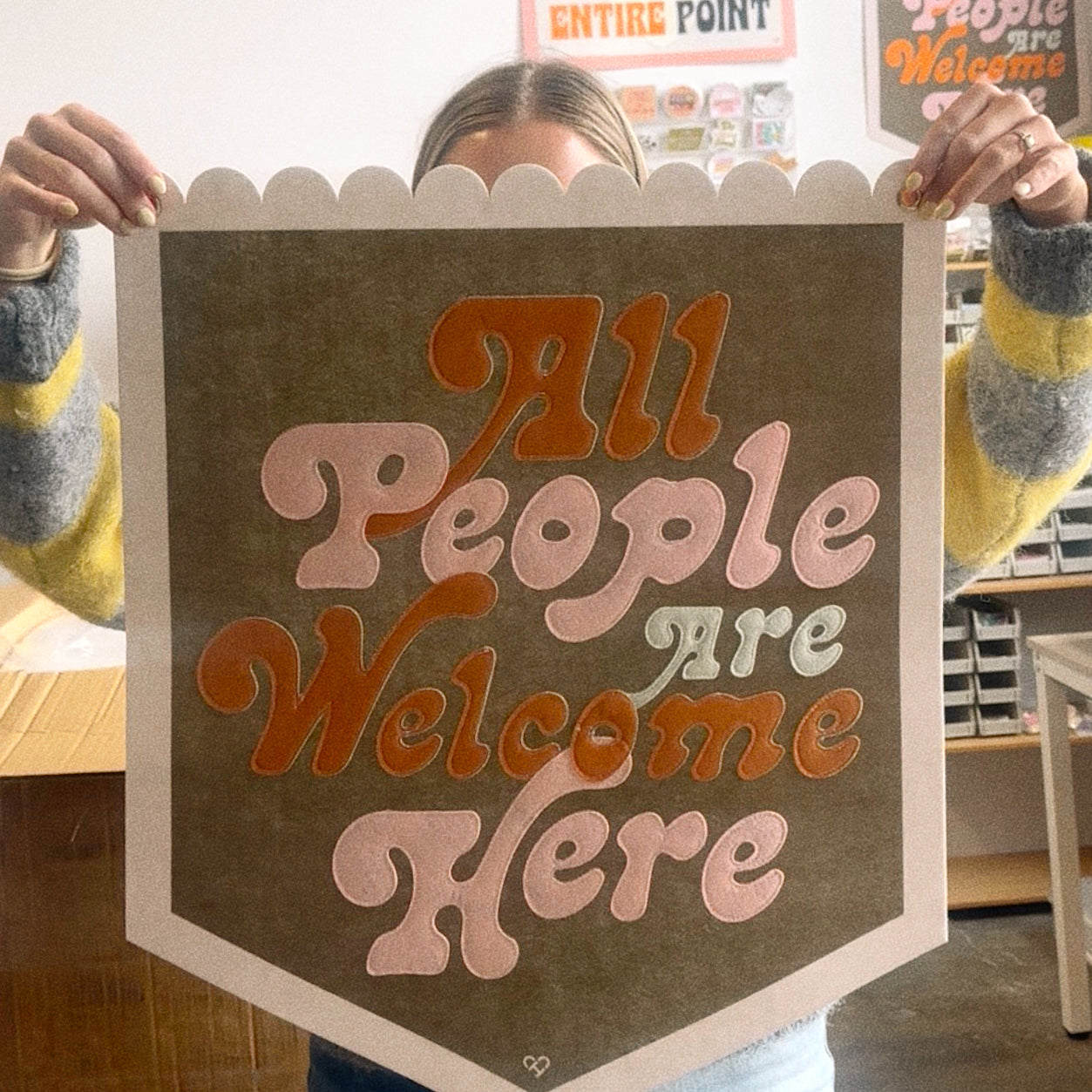 All People Are Welcome Here Pennant, olive scalloped felt banner with pink and orange retro lettering held up