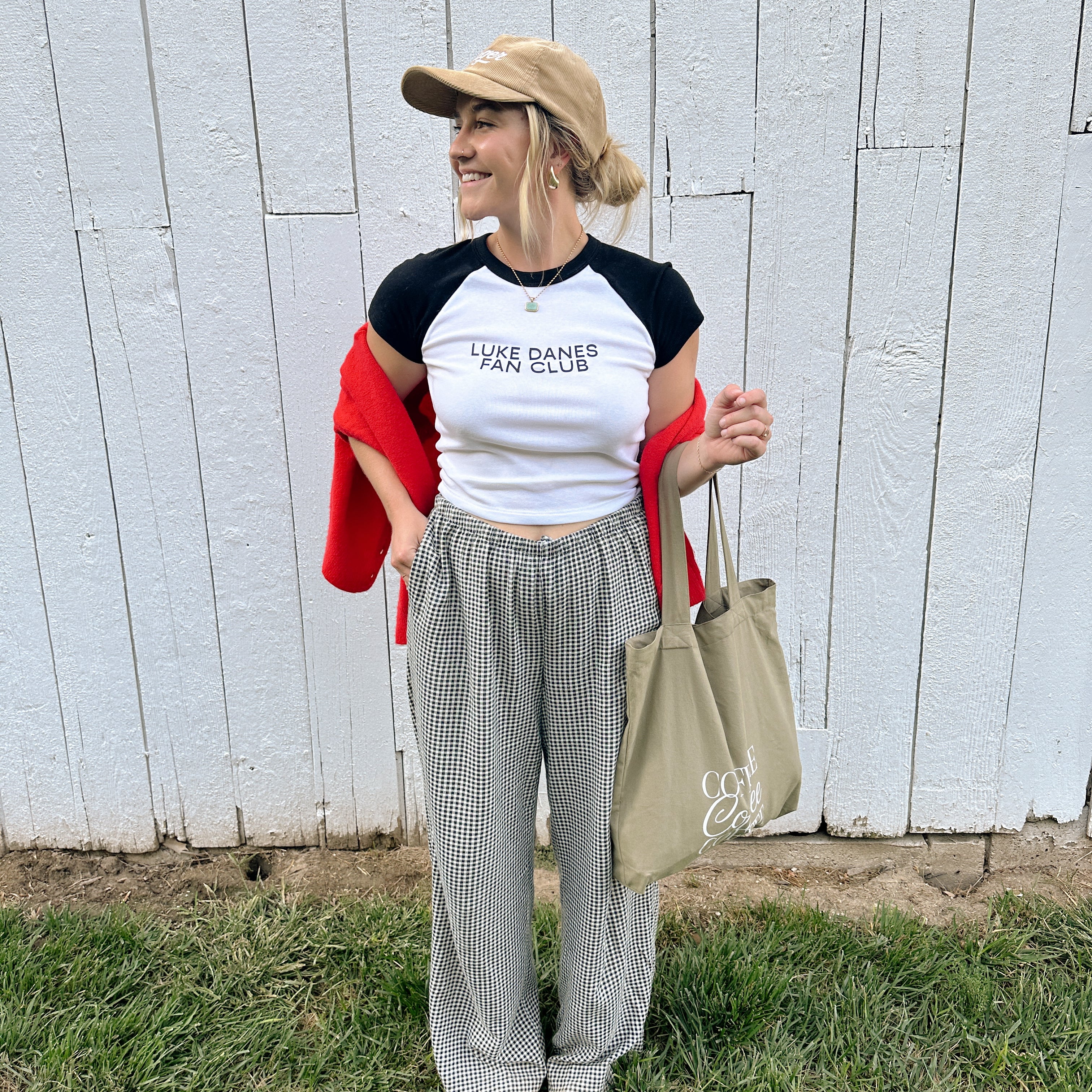 Luke Danes Fan Club Ribbed Tee worn by a model with a tan cap, red sweater and gingham pants by a white barn wall