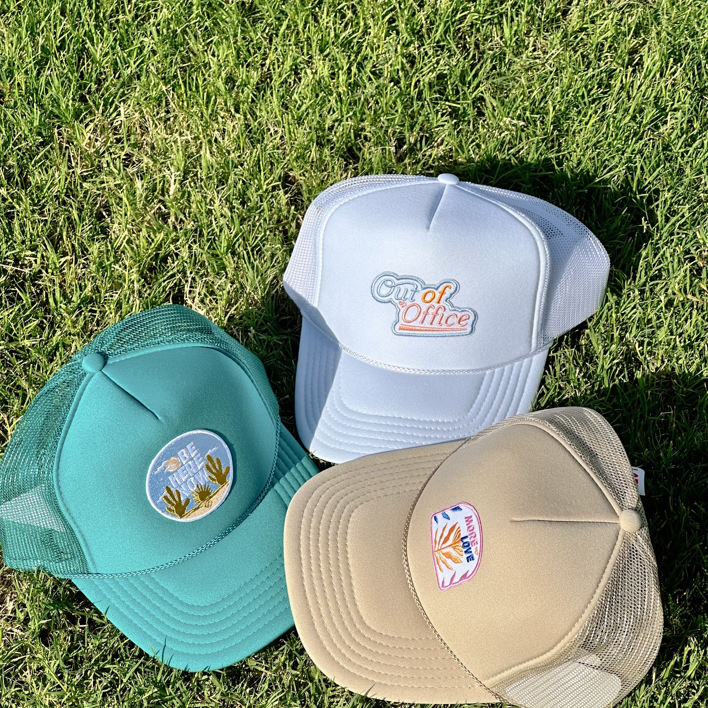 Be Here Now Trucker Hat in teal shown on grass with white Out of Office and tan More Love trucker hats