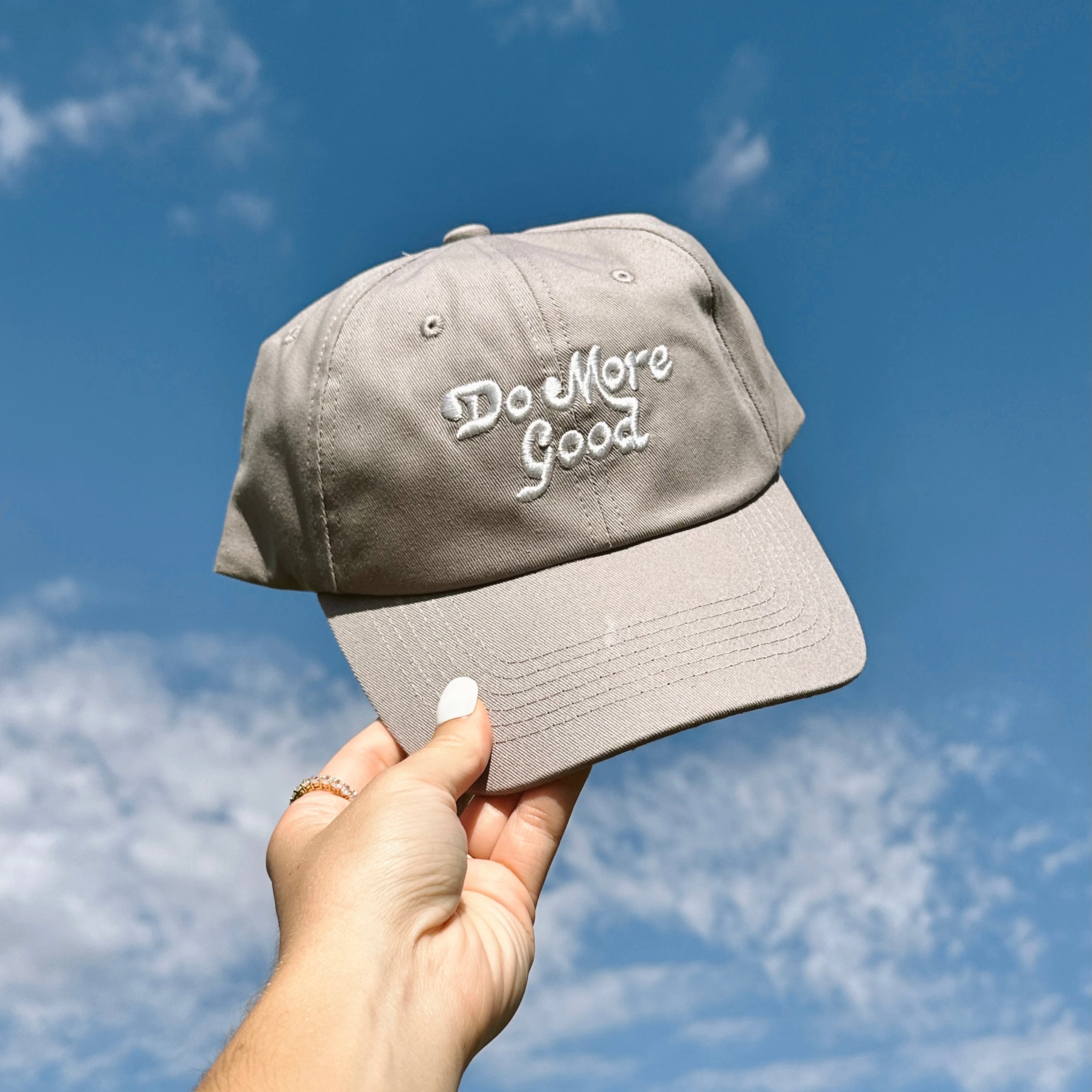 Do More Good Hat, gray baseball cap with white embroidered Do More Good script held up against a blue sky