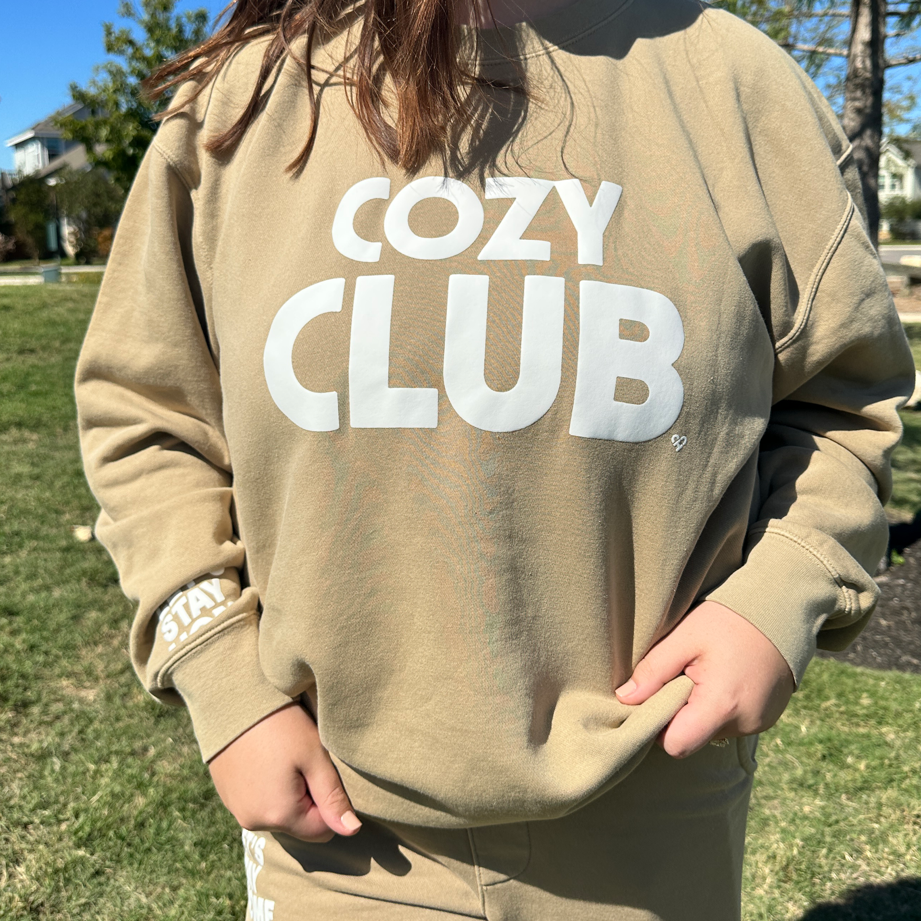 Cozy Club Crew Neck in sandstone with white Cozy Club lettering, worn by a model with matching sweatpants on grass