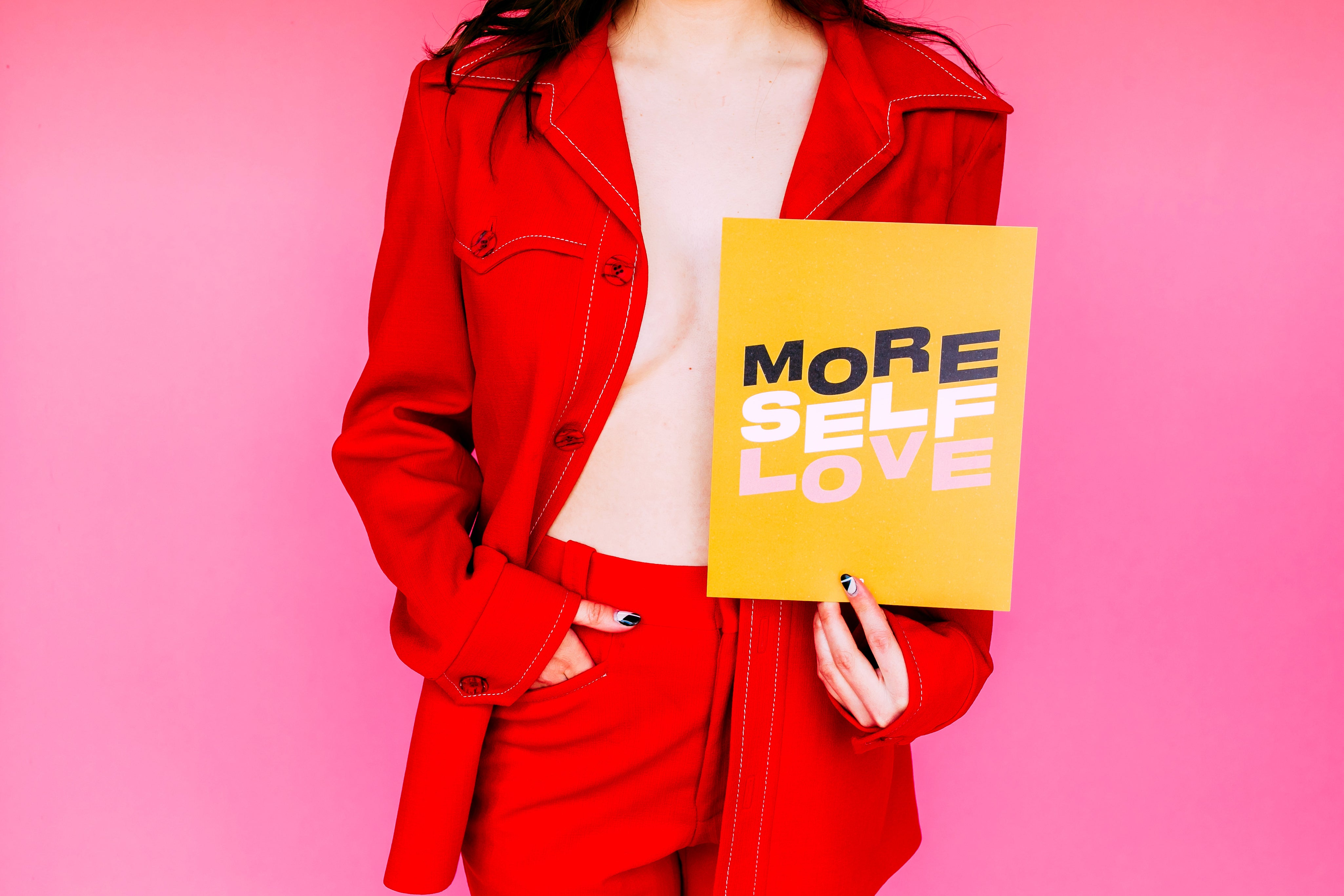 More Self Love Print held by a model in a red suit against a pink wall, mustard print with wavy letters