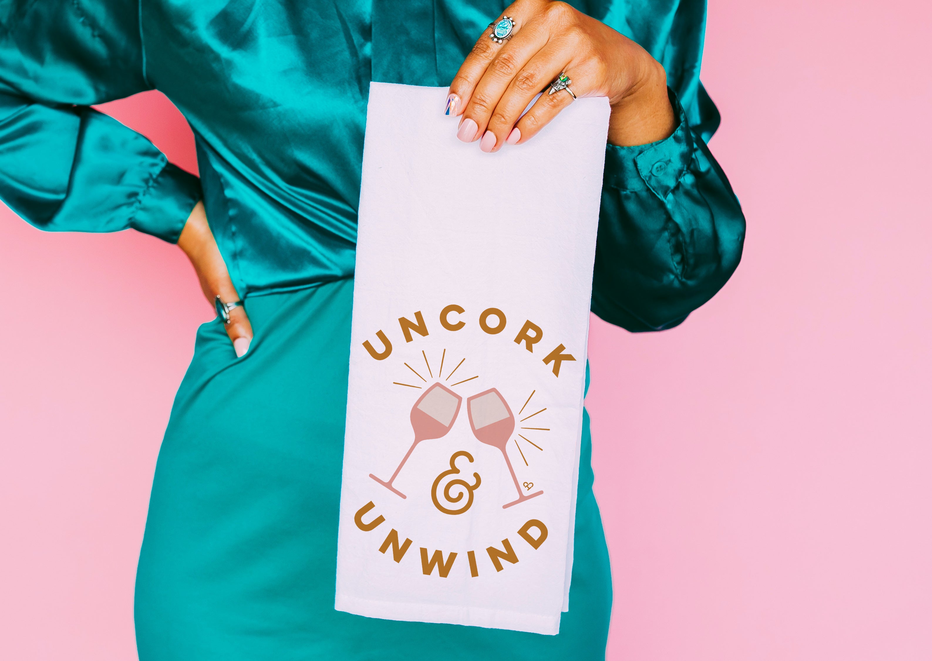 Uncork & Unwind Towel, white tea towel with pink wine glasses and gold text, held by a model in teal satin