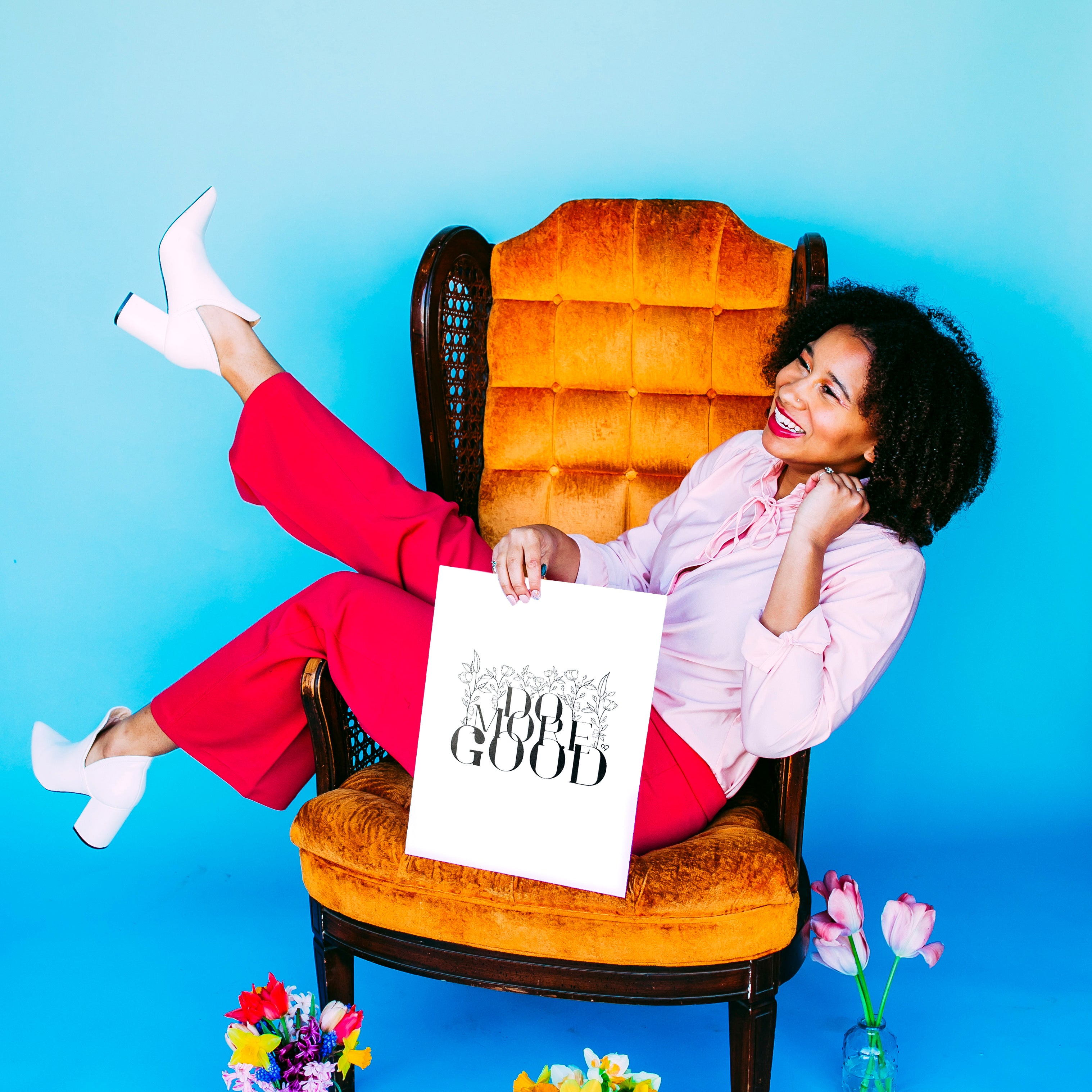 Do More Good White Print held by a smiling model on an orange velvet chair, blue backdrop with tulips
