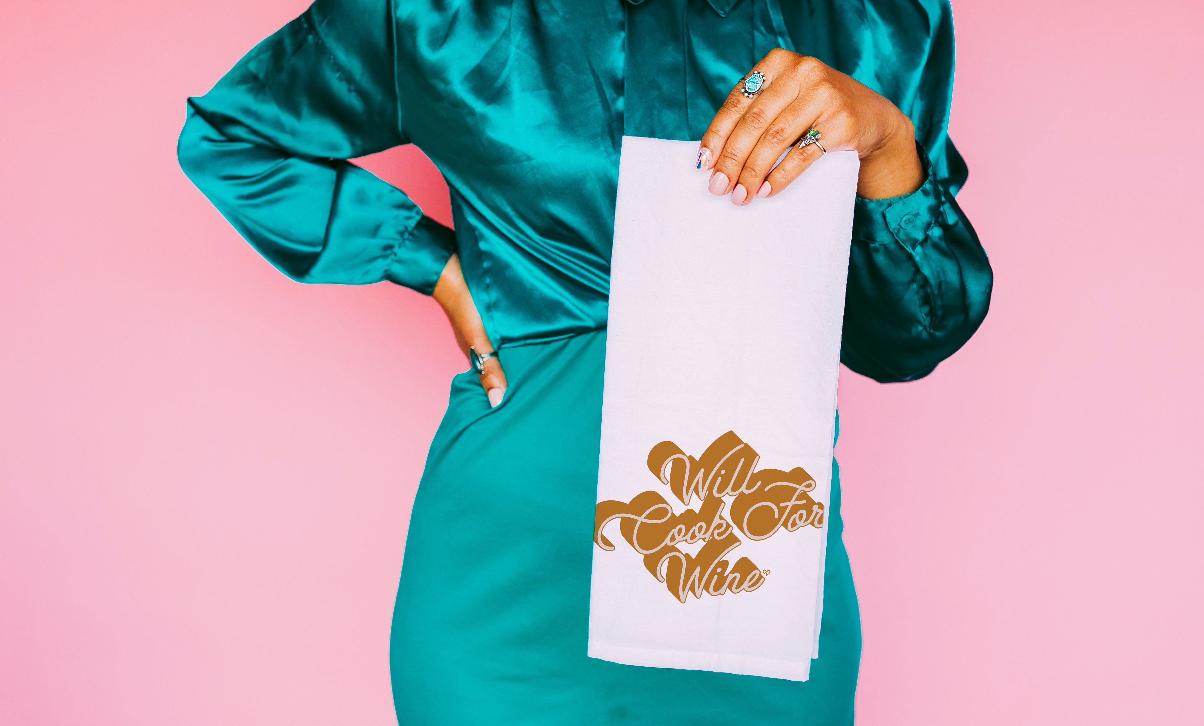 Will Cook For Wine Towel, white tea towel with gold script held by a model in teal satin, pink backdrop