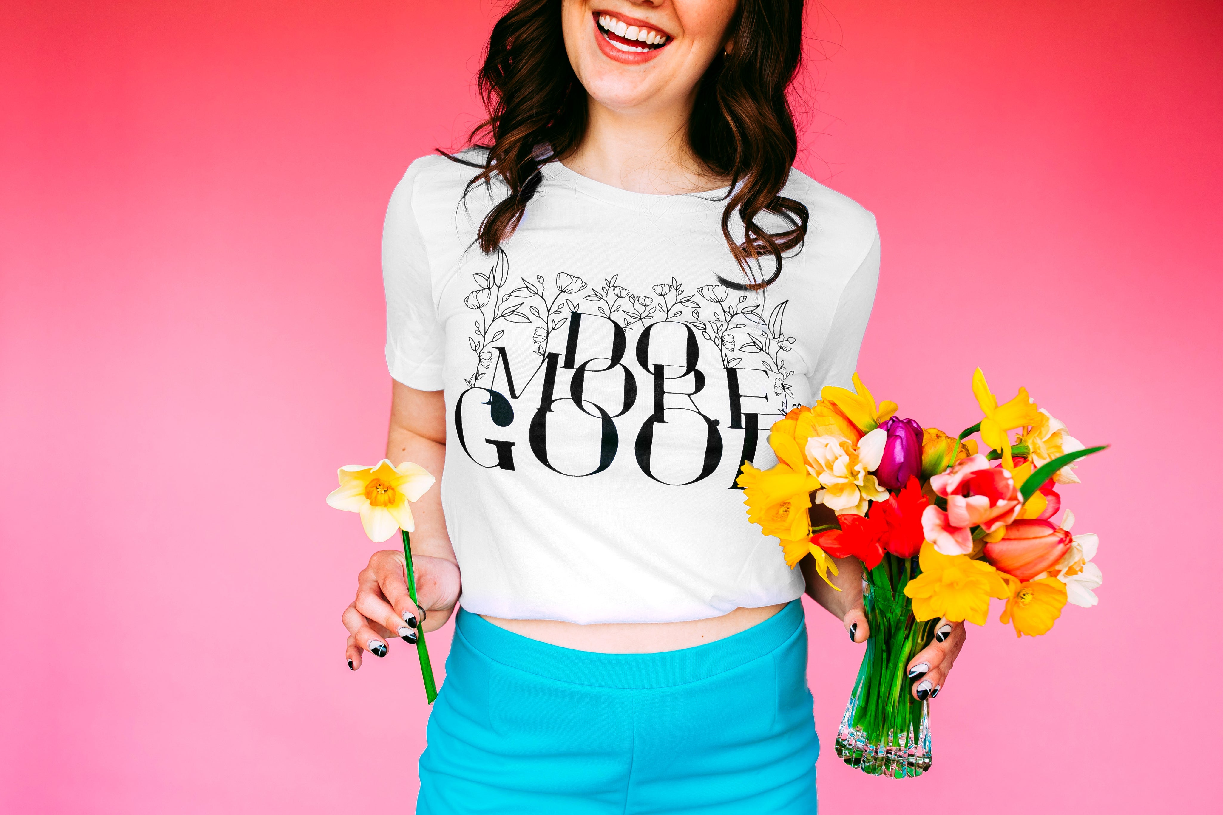 Do More Good Tee worn by a smiling model holding tulips and daffodils against a pink backdrop