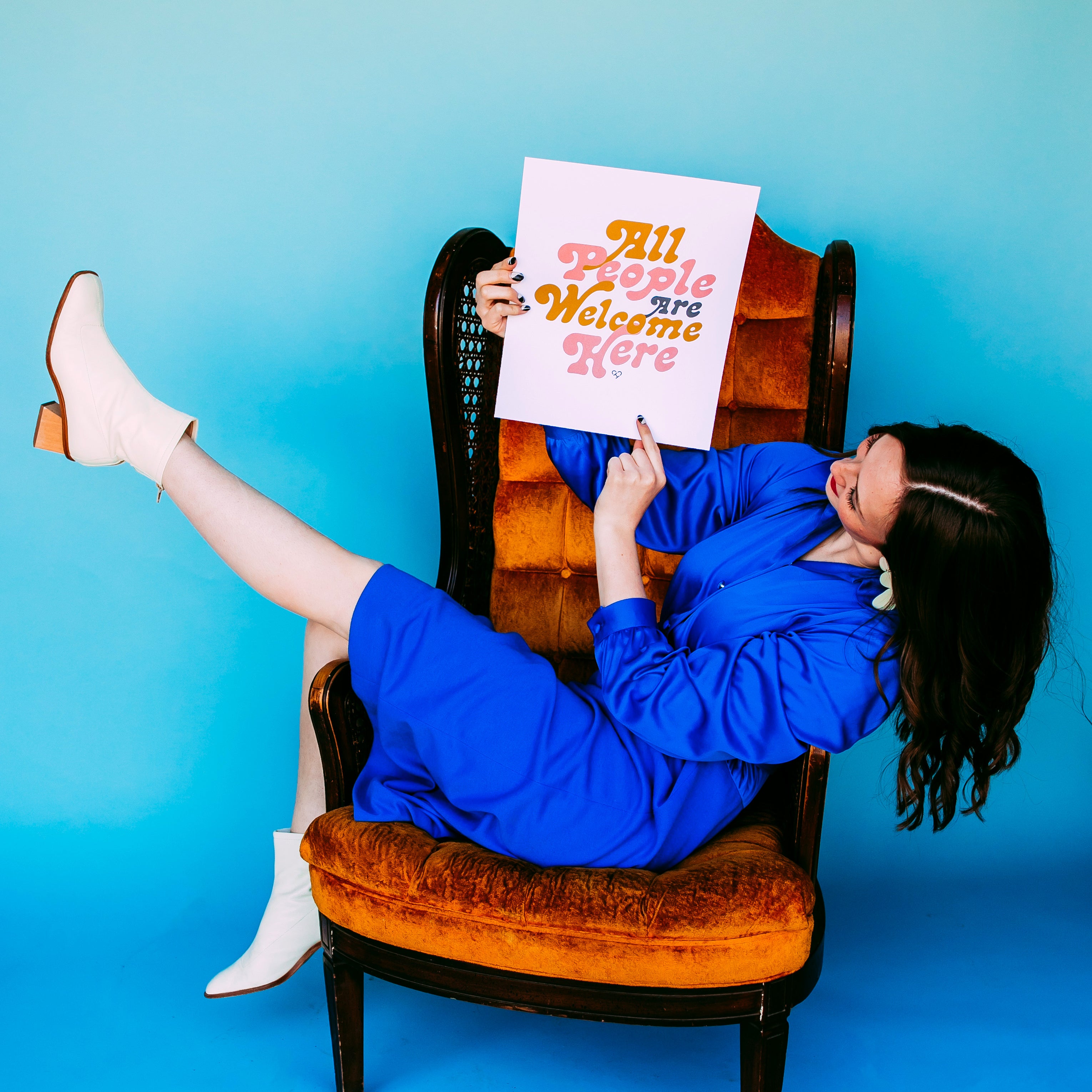 All People Are Welcome Here Print held by a model in blue lounging on an orange velvet chair, blue backdrop