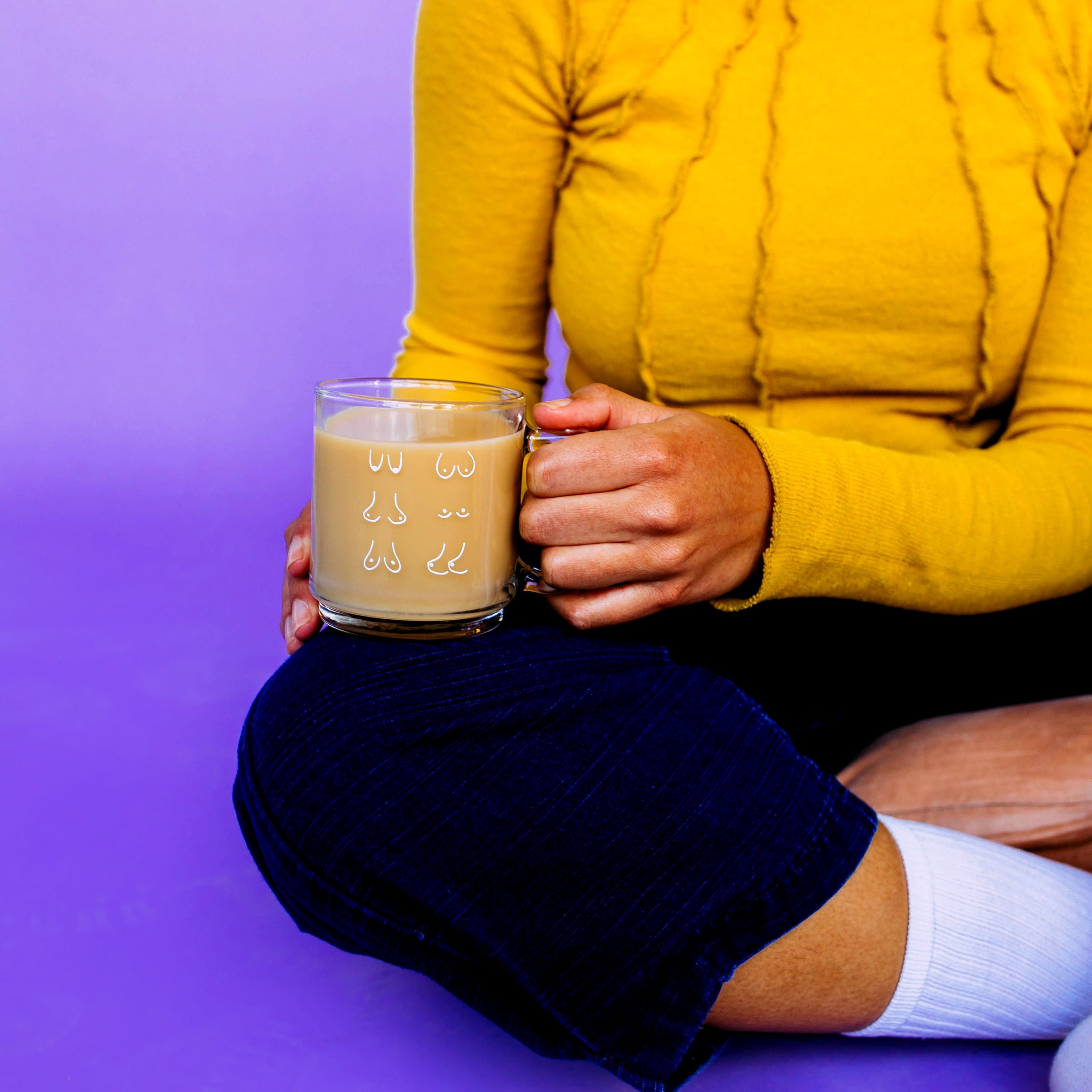Glass Boob Mug, clear glass mug of coffee with line drawn breasts held by a model in yellow, purple backdrop